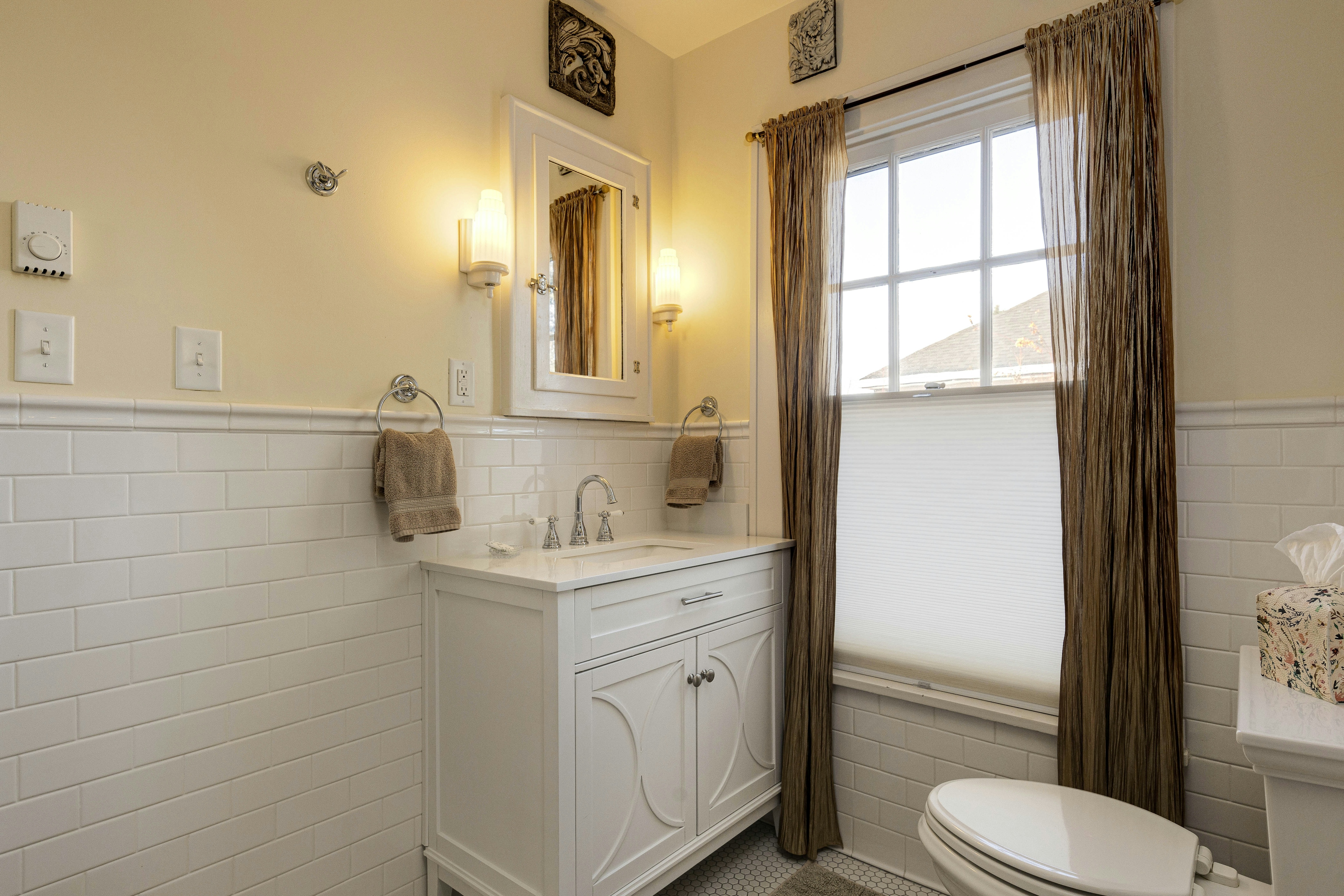 A bathroom with a white vanity, a wall-mounted mirror, two sconces, a window with curtains, and beige walls with white tile wainscoting.