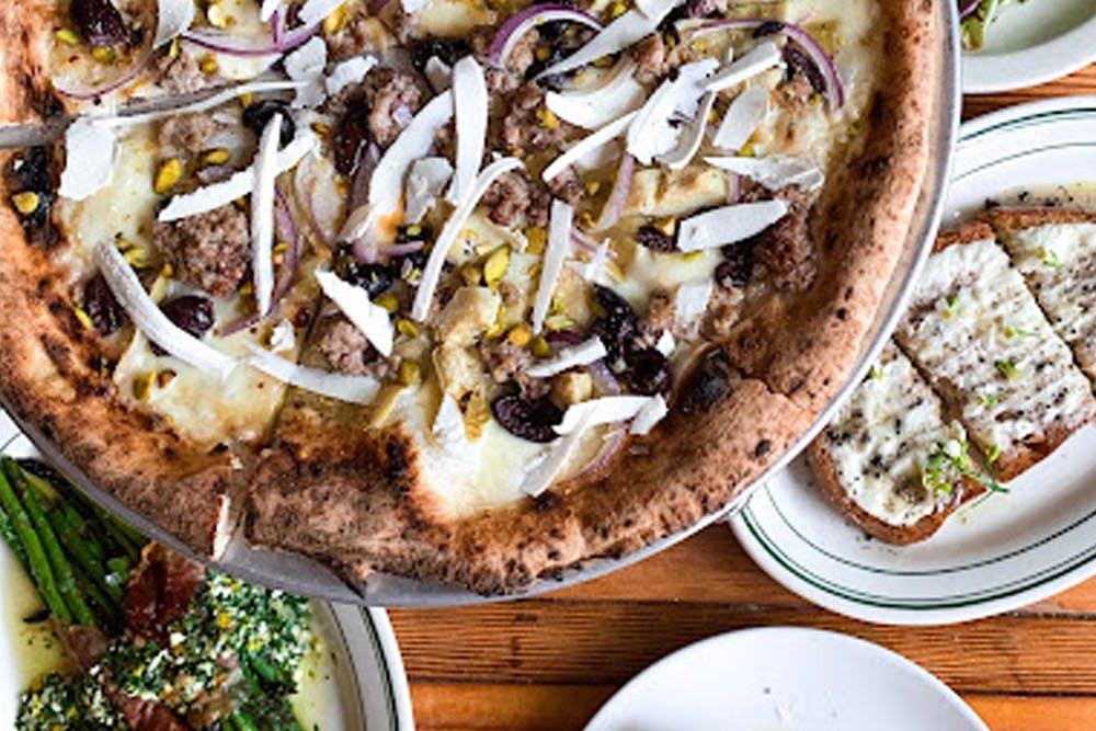 A wood-fired pizza topped with sausage, olives, onions, and cheese, accompanied by side dishes including garlic bread and asparagus.