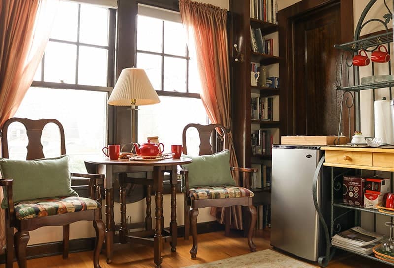 A cozy sitting area with two cushioned chairs and a small wooden table set with a red teapot and cups, positioned by a window with sheer curtains. A small fridge and a shelving unit holding kitchen items are in the corner of the room.