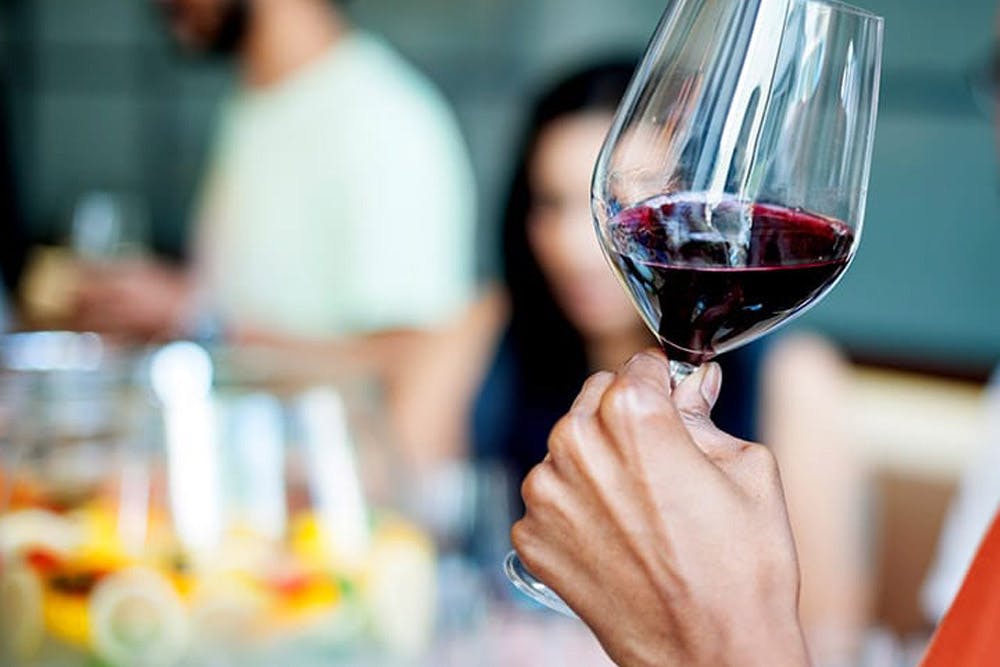 A person holding a glass of red wine, with a blurred background of people and a table of food.