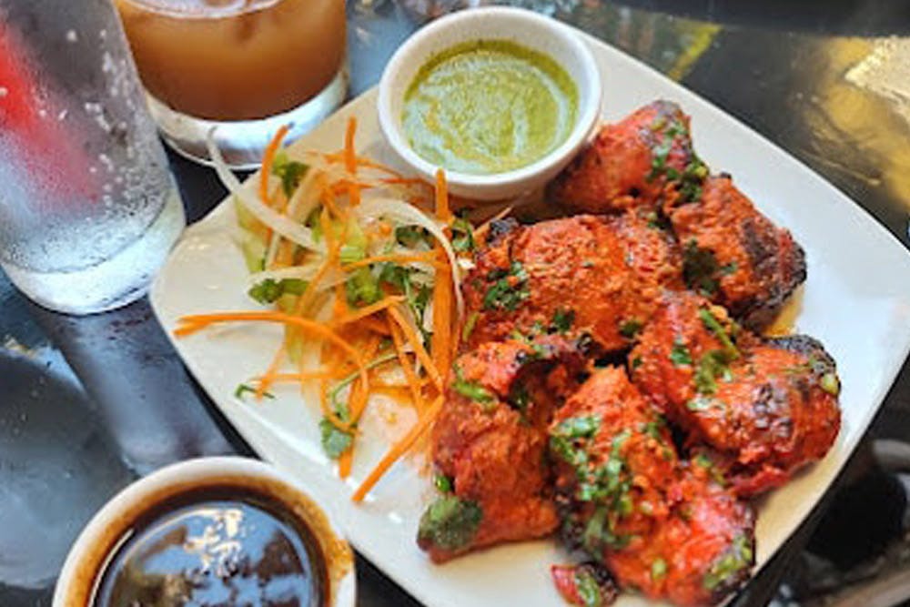 A plate of tandoori chicken with a side of mixed vegetables, green chutney, and a glass of water and a drink.