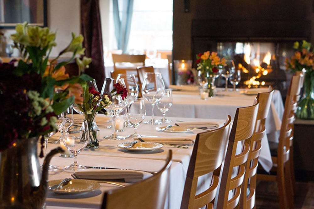 An elegant dining table set with white linens, flowers, and glassware in a restaurant with a cozy fireplace in the background.