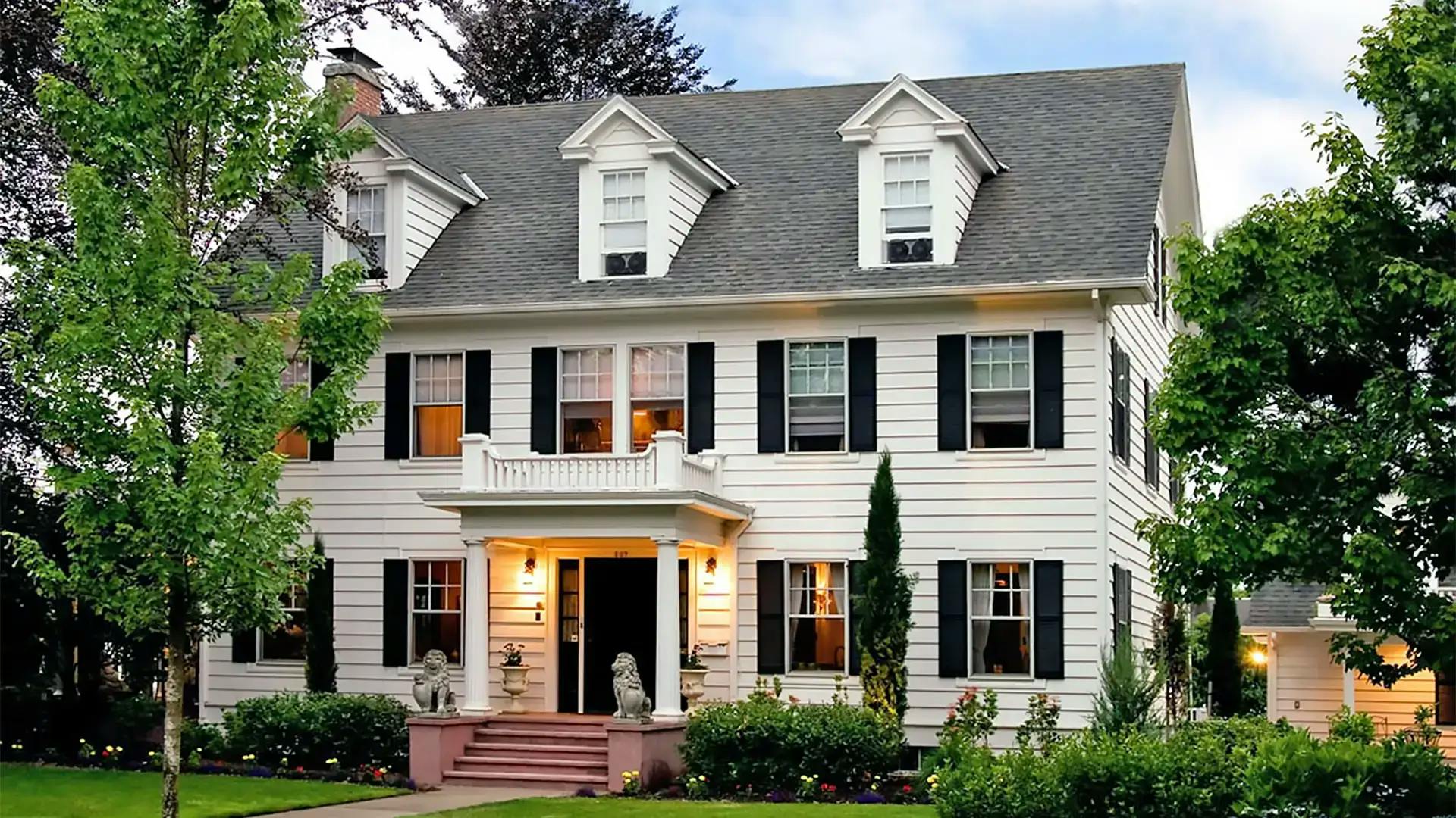 A large, classic white house with black shutters and dormer windows, featuring a covered entrance with columns, surrounded by manicured bushes and trees.