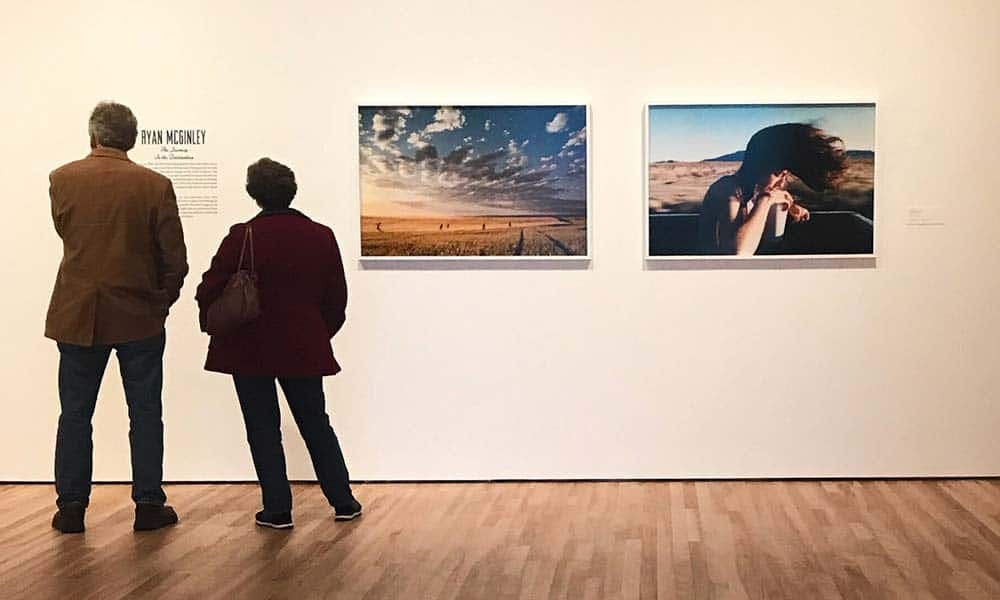 Two people standing in an art gallery, viewing photographs on a white wall; one photo shows a vast landscape with a dramatic sky, and the other features a close-up of a person with windblown hair.