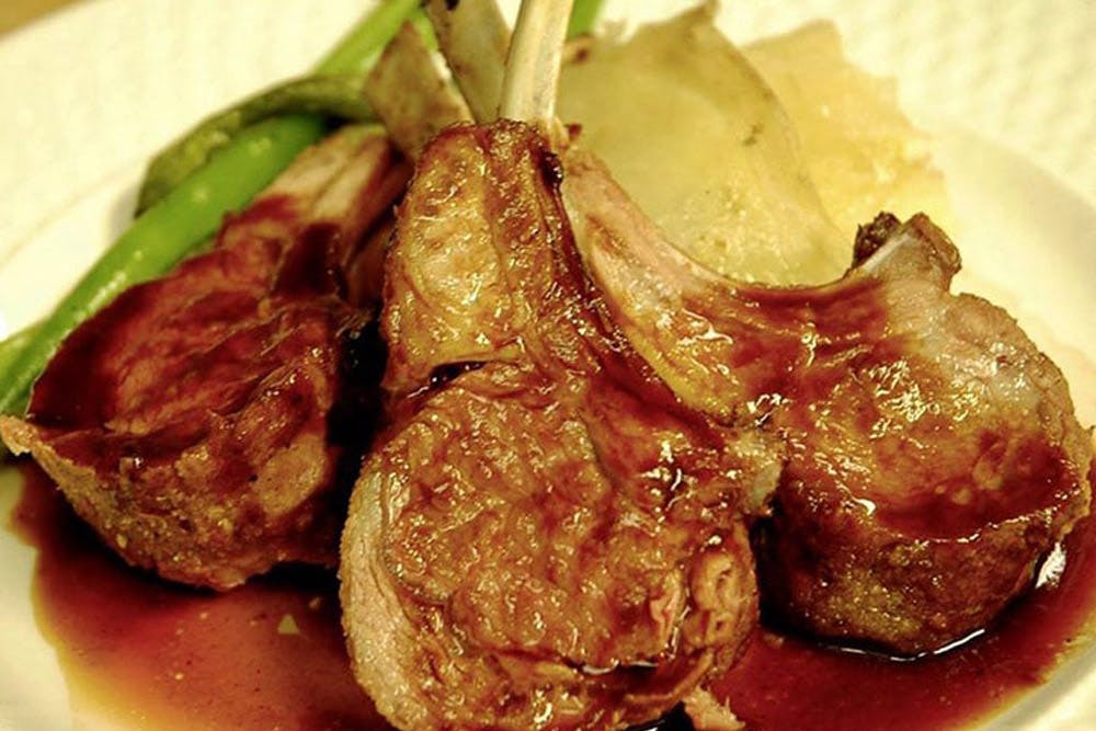 A plate of succulent lamb chops with a rich sauce, served with green vegetables and a side of potatoes.