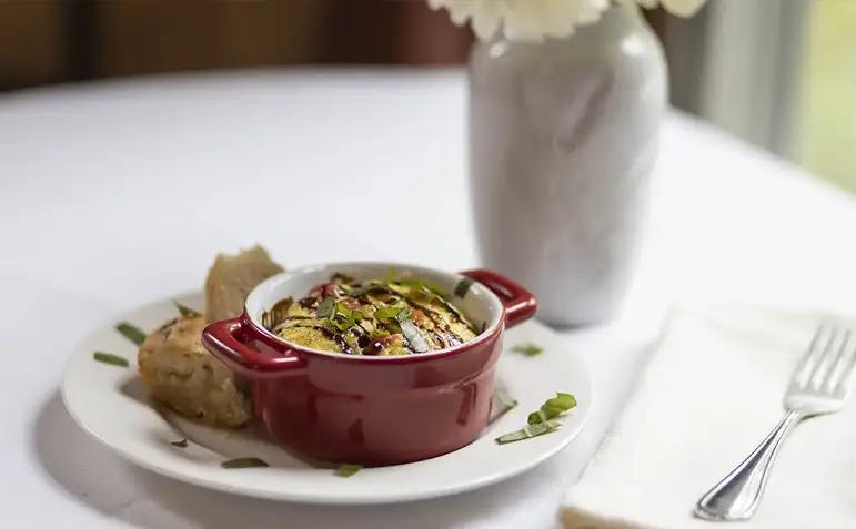 A red ceramic dish filled with a baked frittata, garnished with fresh herbs, sits on a white plate with a side of bread. A white napkin with a fork is placed nearby, and a white vase with flowers adds a decorative touch in the background.