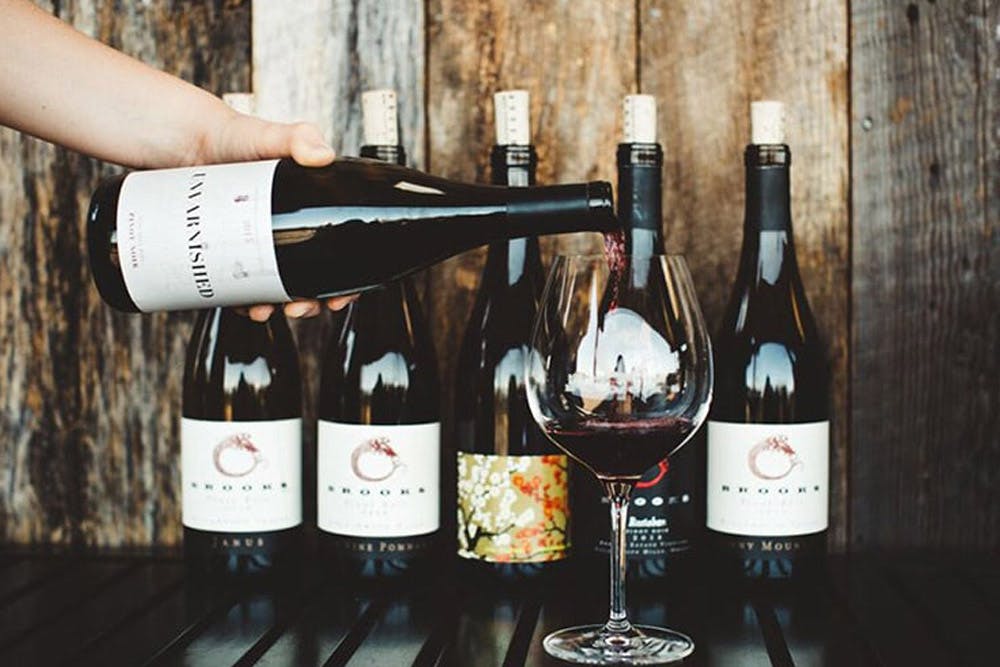 A hand pouring red wine from a bottle labeled "Unfinshed" into a glass, with several other bottles of wine lined up on a wooden surface in the background.