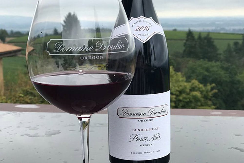 A glass of red wine and a bottle of Domaine Drouhin Oregon Pinot Noir 2016 set on a table, with a scenic vineyard and countryside view in the background.