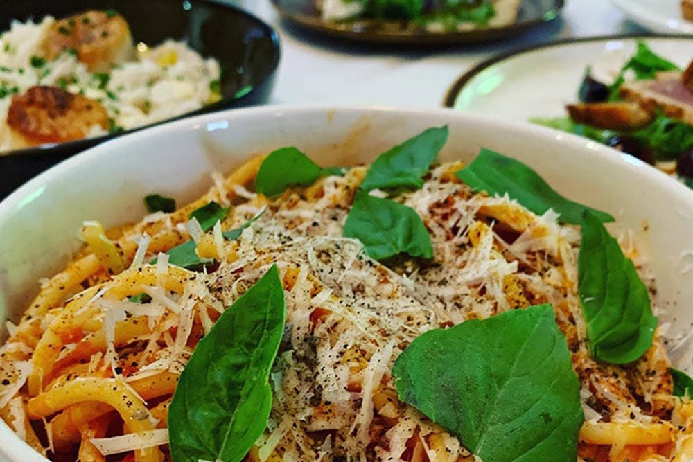 A bowl of pasta garnished with fresh basil leaves and grated cheese, with plates of risotto and other dishes in the background.