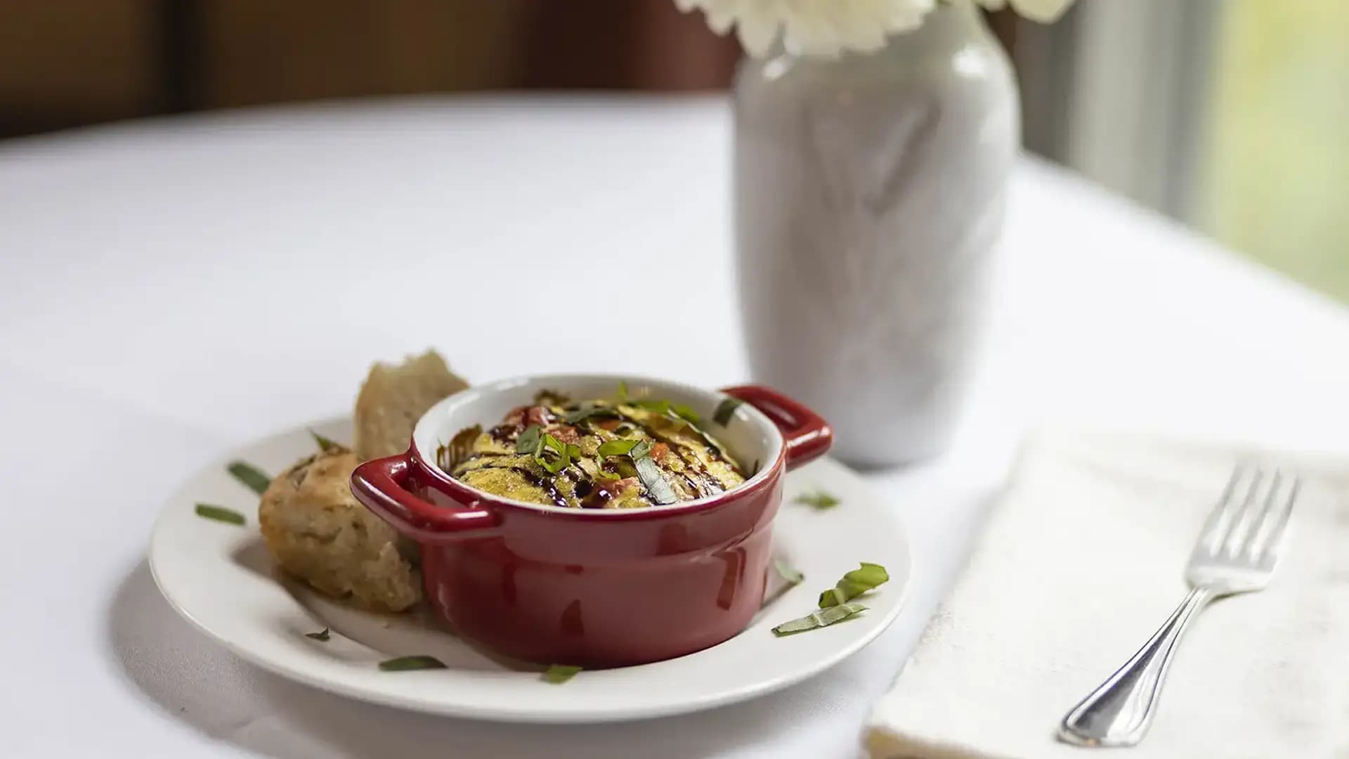 A small red casserole dish filled with a savory baked dish, garnished with herbs, served with bread on a white plate, placed on a white tablecloth with a fork and a vase of flowers nearby.