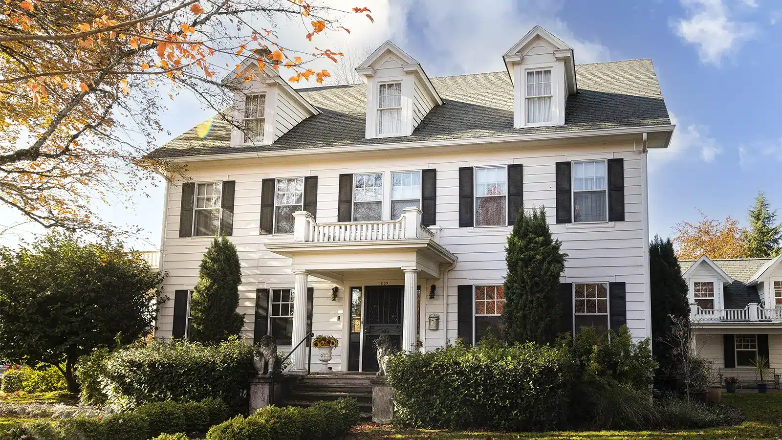 A large, historic white house with black shutters, dormer windows, and a covered front entrance, surrounded by lush greenery and autumn foliage.