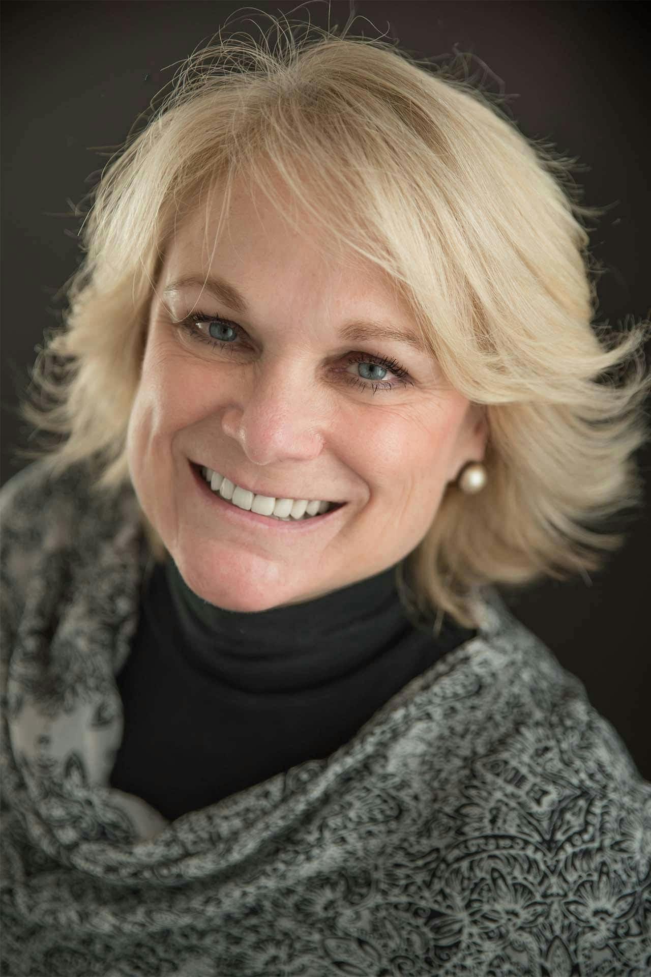 A smiling woman with short blonde hair, wearing a patterned scarf and pearl earrings.