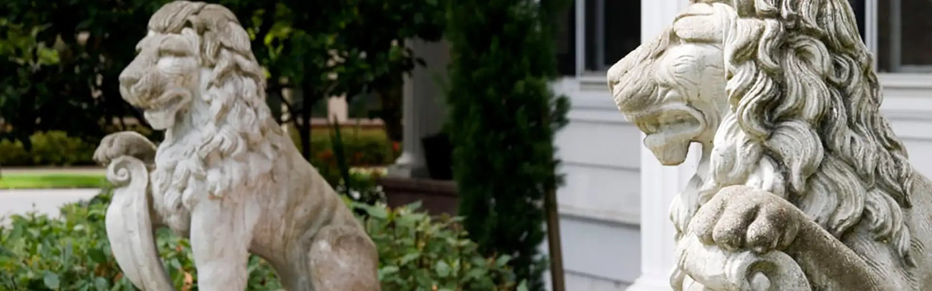 Two stone lion statues in a garden setting, positioned in front of a house, with detailed manes and a regal appearance.