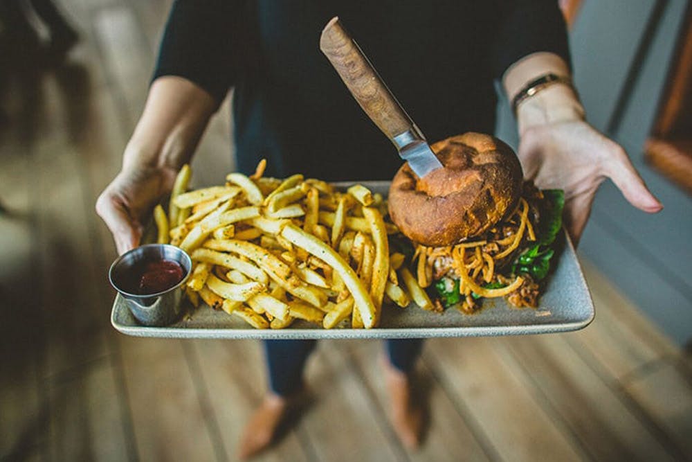 A person holding a plate with a burger topped with fried onions, a knife stuck in the bun, and a side of seasoned fries with ketchup.