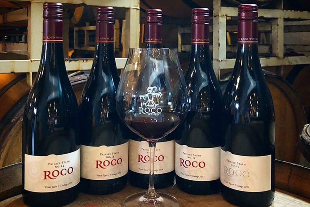 A glass of red wine in front of several bottles of Roco Winery Pinot Noir, arranged on a wooden barrel in a wine cellar.