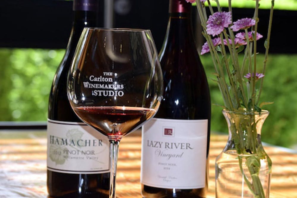 A glass of red wine with "The Carlton Winemakers Studio" logo, placed on a table with two bottles of wine and a vase with flowers.