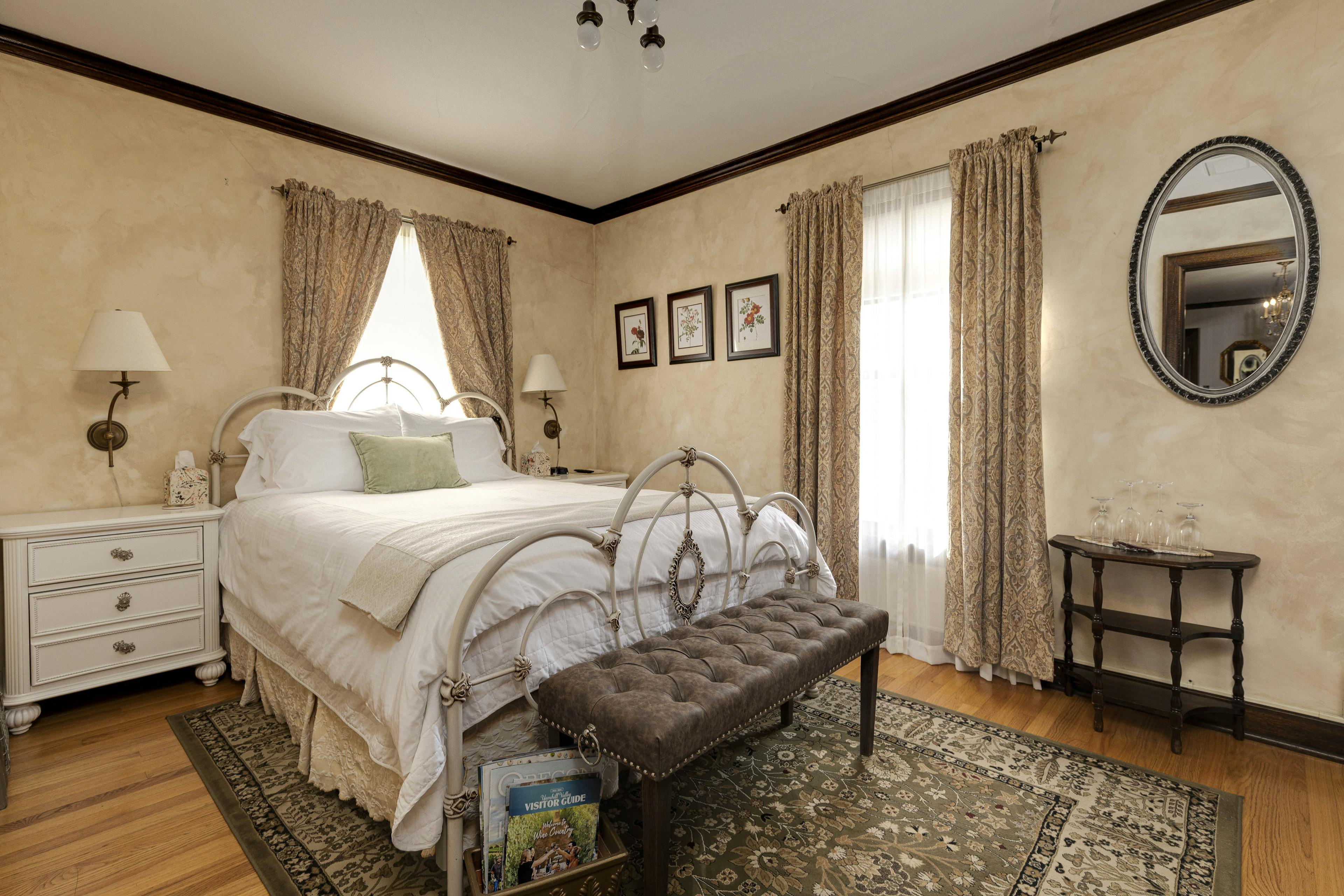 The room has a metal-framed bed with white linens, floral prints on the walls, and elegant curtains. A tufted bench sits at the foot of the bed, and a side table with wine glasses adds a touch of elegance.