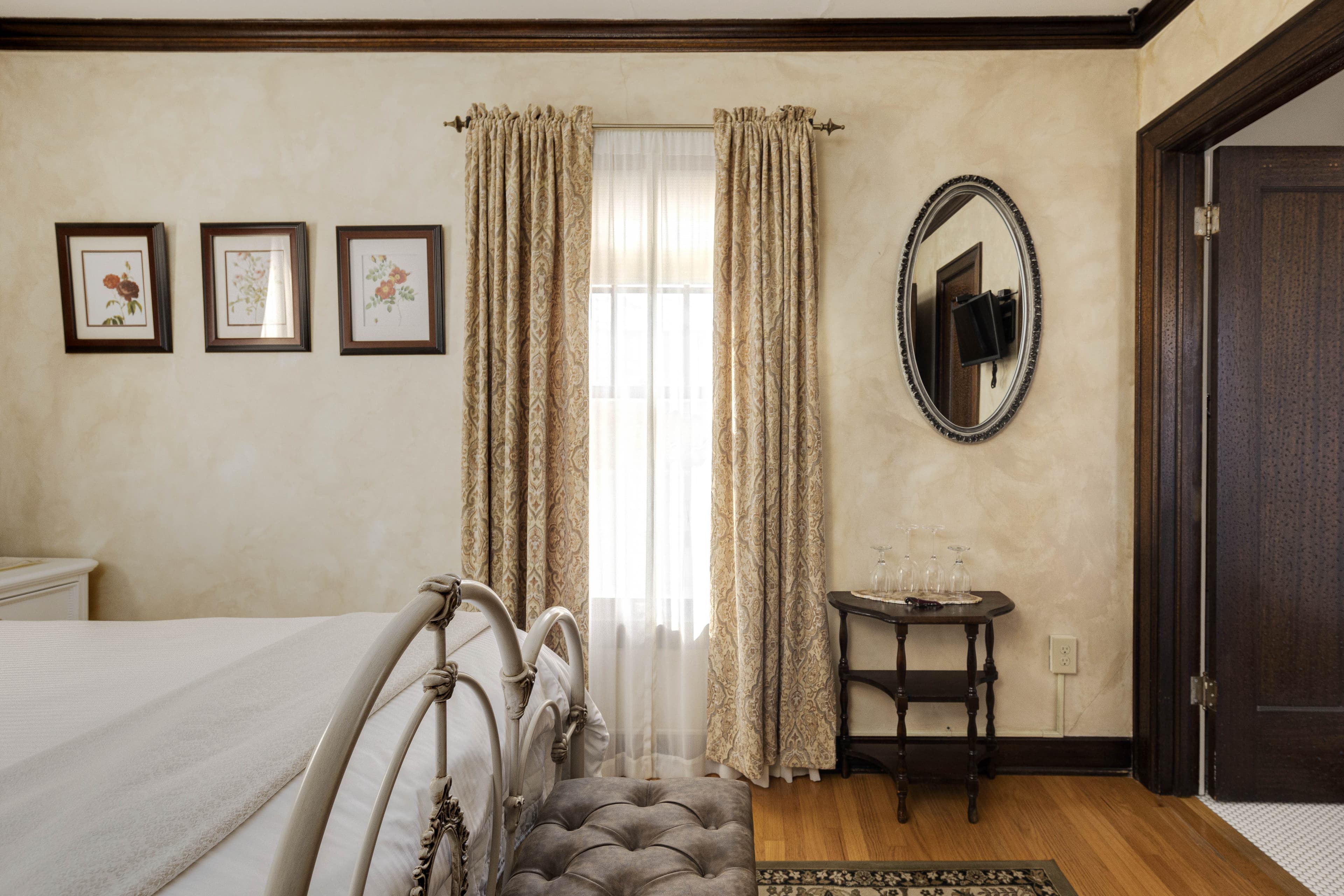 A cozy room featuring a metal-framed bed, floral prints on the wall, and patterned curtains. An oval mirror hangs above a wooden side table with wine glasses.