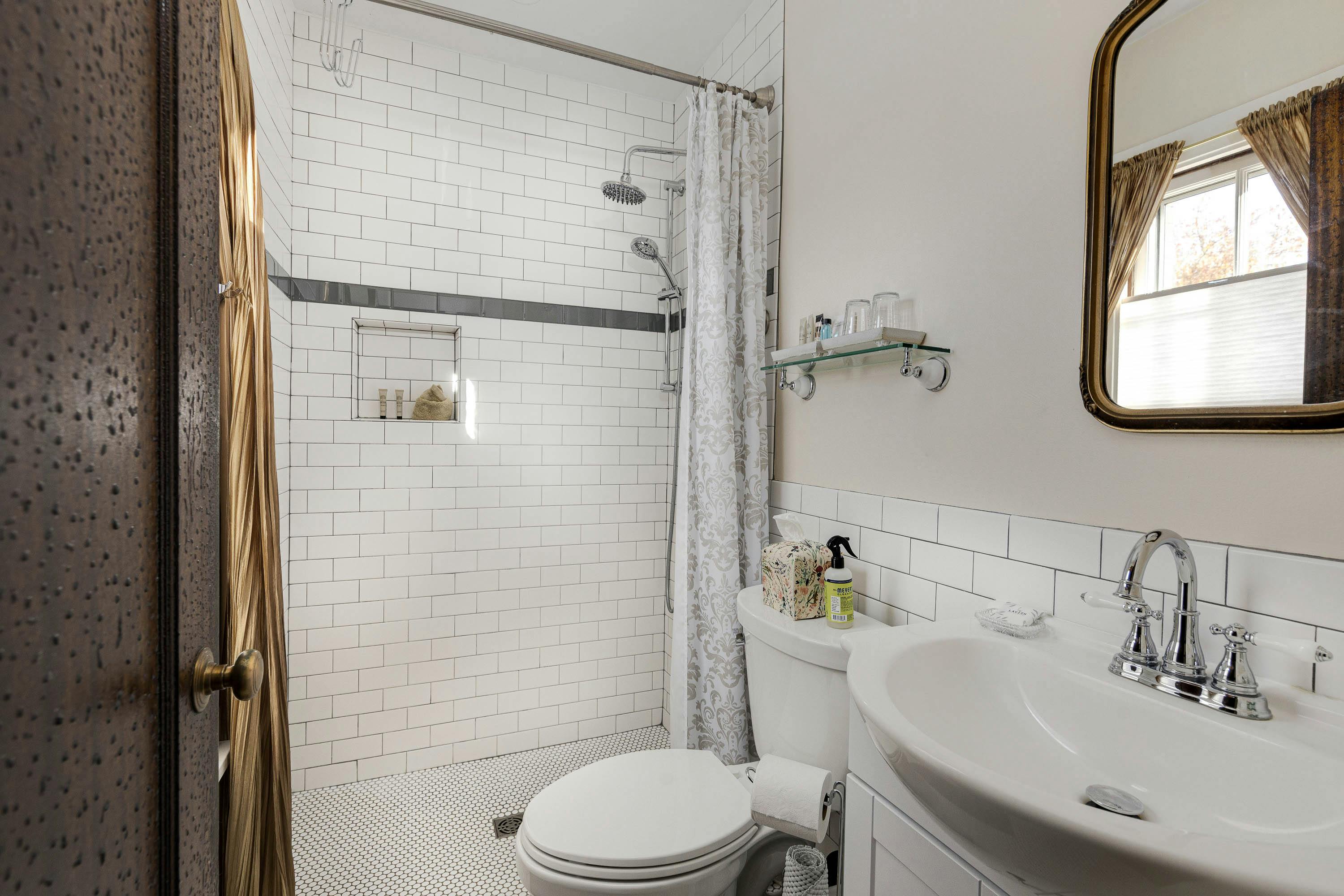 A bright bathroom featuring white subway tiles, a shower area with a curtain, a toilet, a sink, a glass shelf with toiletries, and a mirror.