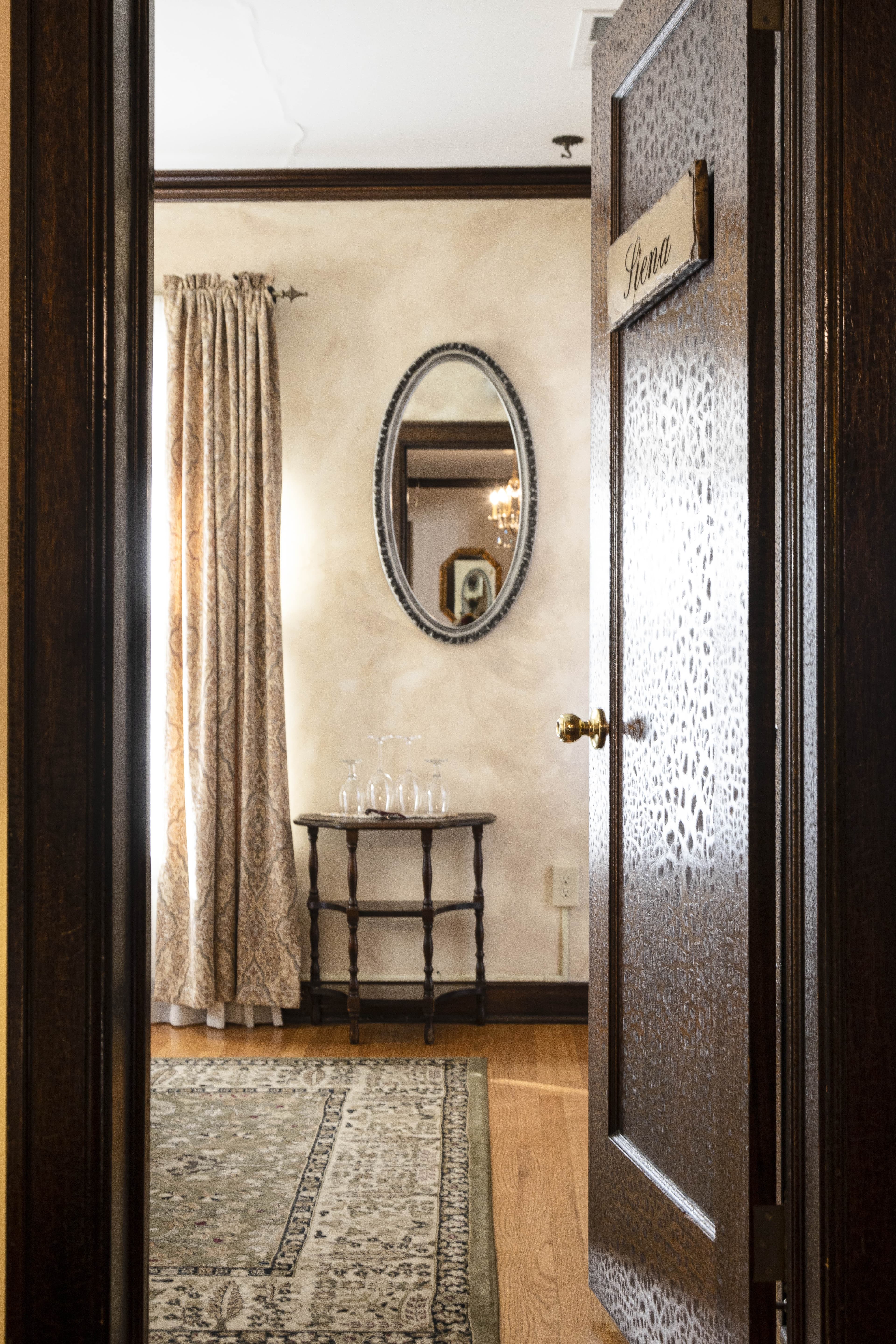 An open door labeled "Siena" leads into a room with a mirror, patterned curtains, a small table with glasses, and a patterned rug.