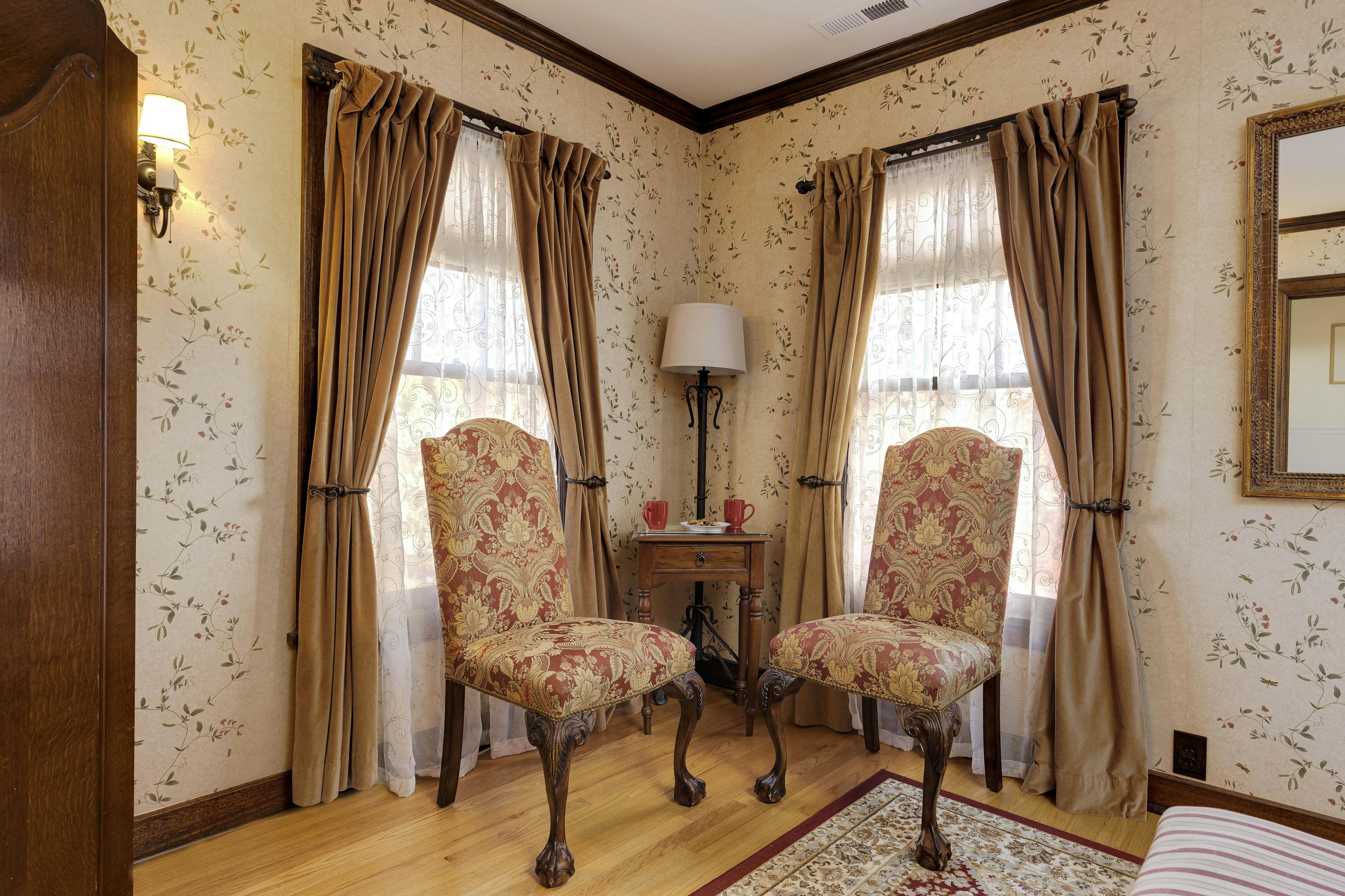 A cozy corner of a room with two ornate, upholstered chairs placed around a small wooden table with a lamp, flanked by windows draped with sheer and heavy curtains, all against wallpaper featuring a delicate floral pattern.