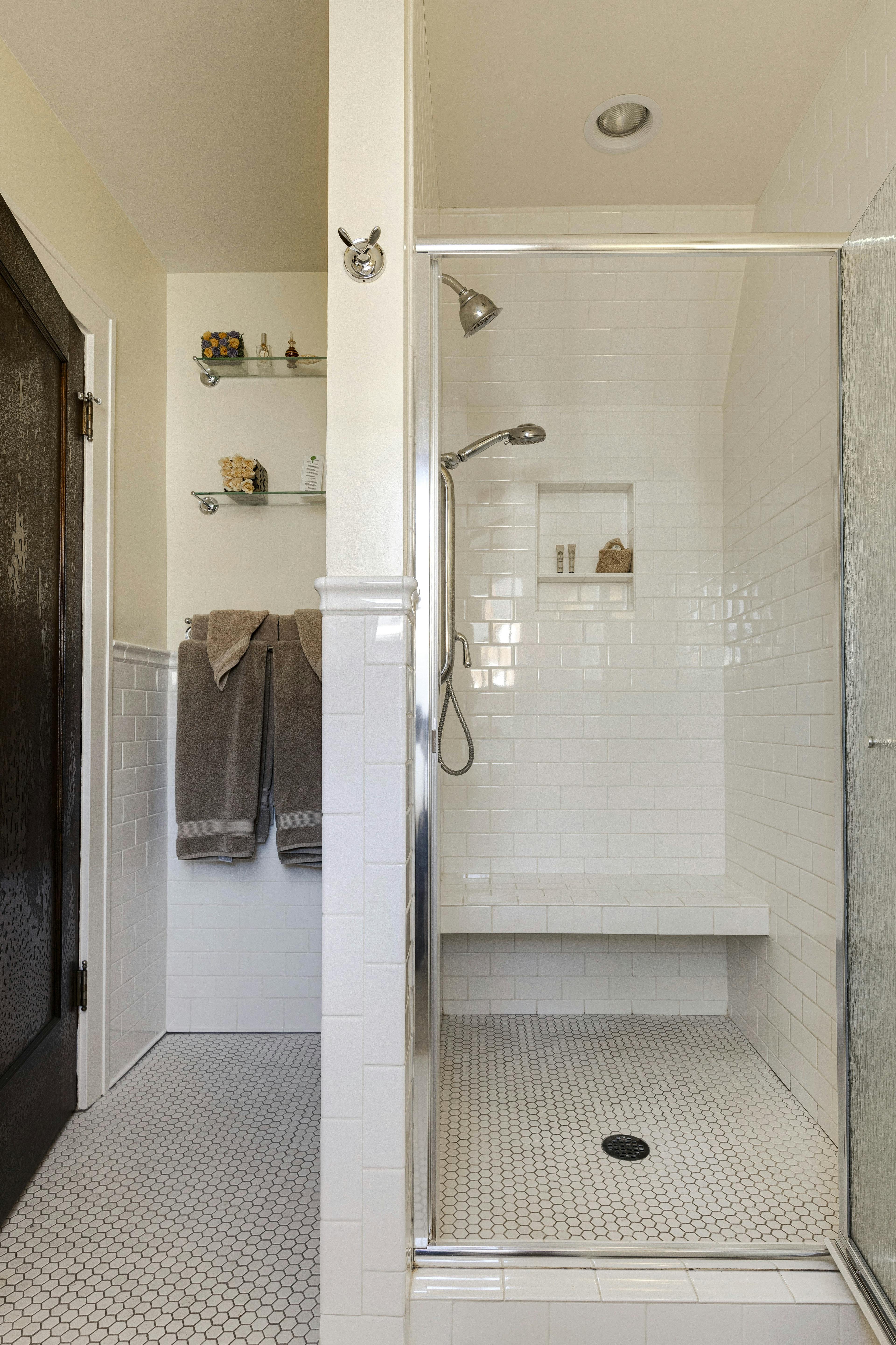 A tiled bathroom with a walk-in shower, glass door, white subway tiles, a built-in bench, and shelves holding folded towels and toiletries.