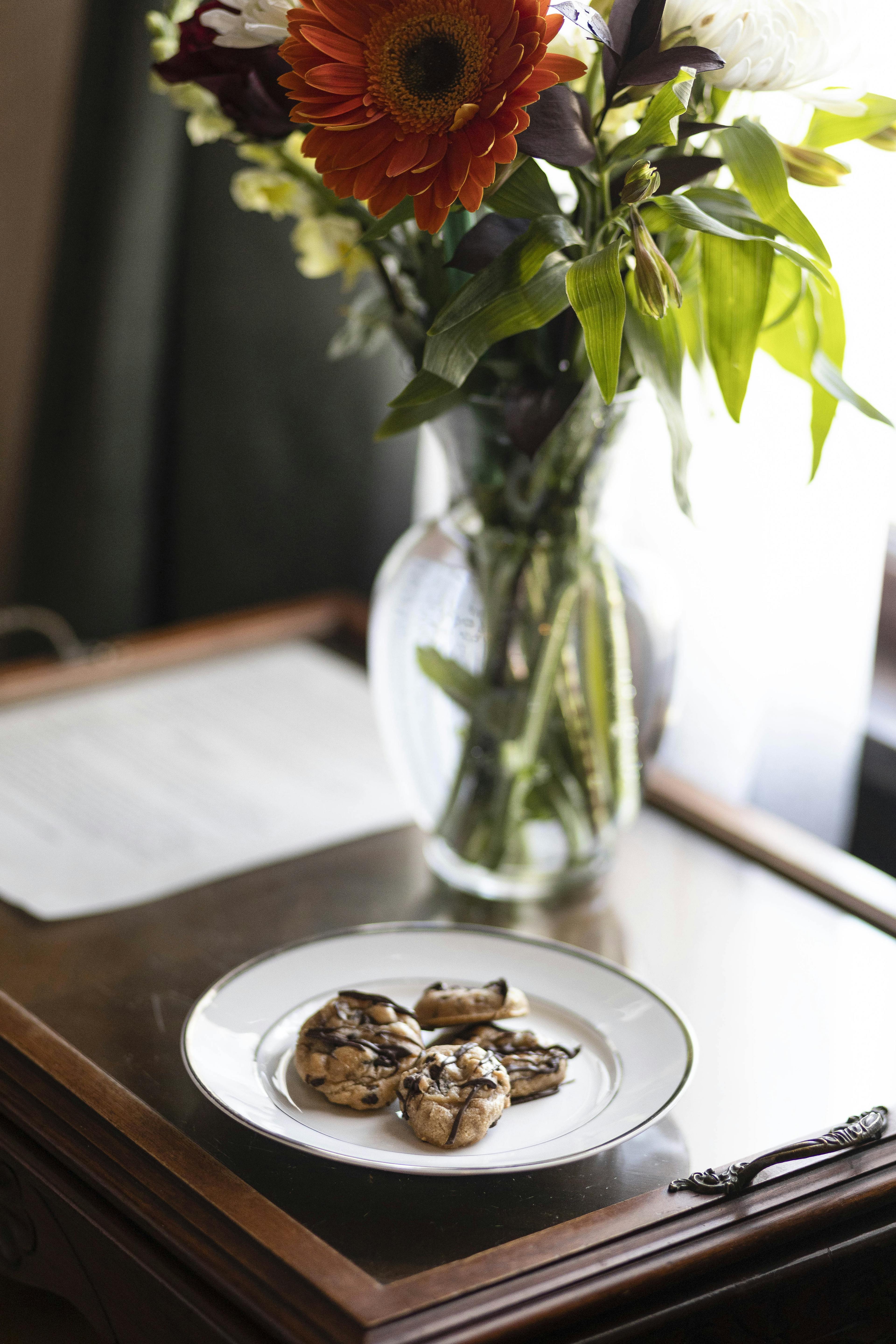 A plate of chocolate-drizzled cookies on a wooden table next to a vase of flowers in the Napoli Guest Room.