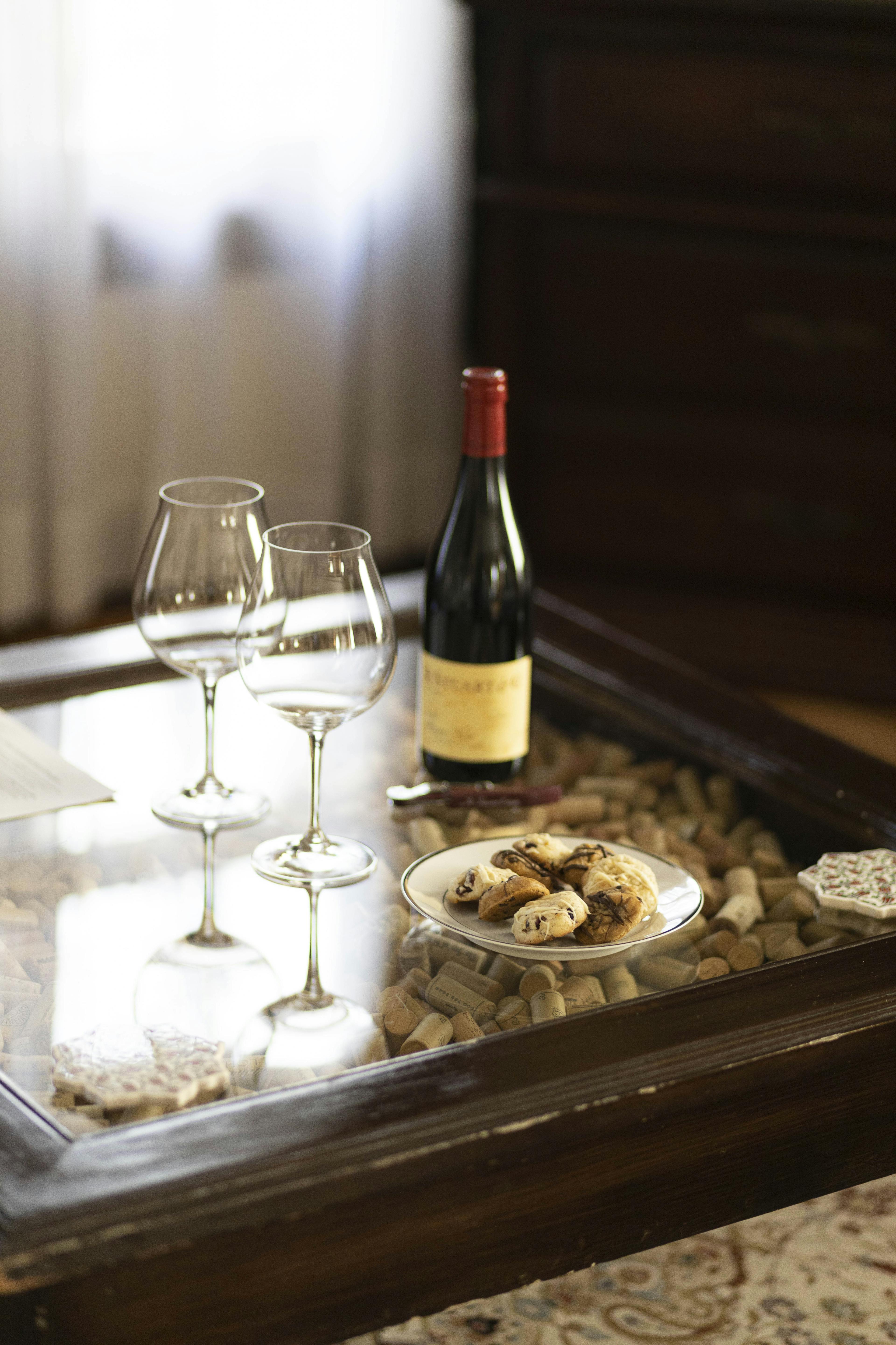 A close-up of a glass-topped table with two wine glasses, a bottle of red wine, and a plate of cookies, set against a blurred background.