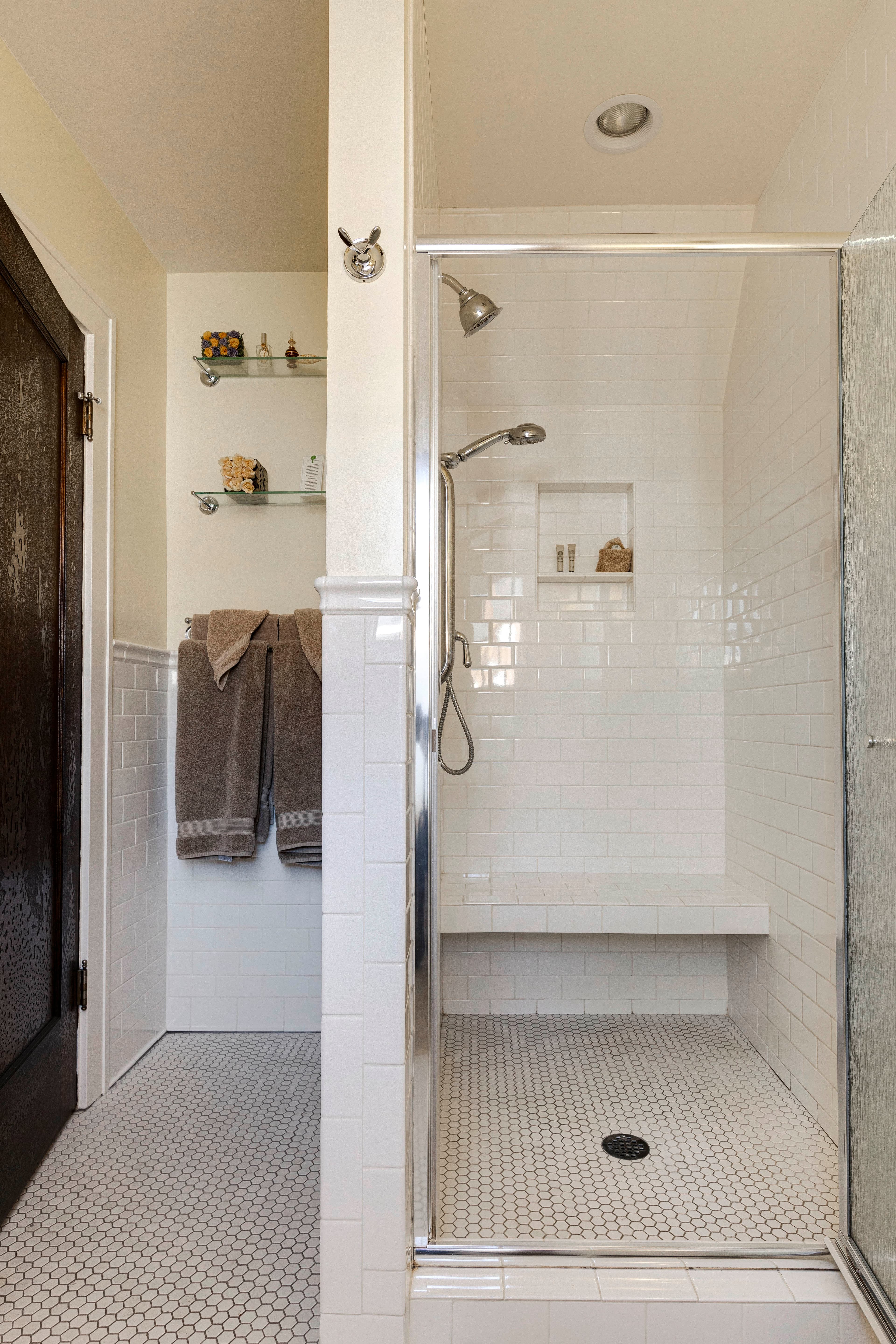A tiled bathroom with a walk-in shower, glass door, white subway tiles, a built-in bench, and shelves holding folded towels and toiletries.
