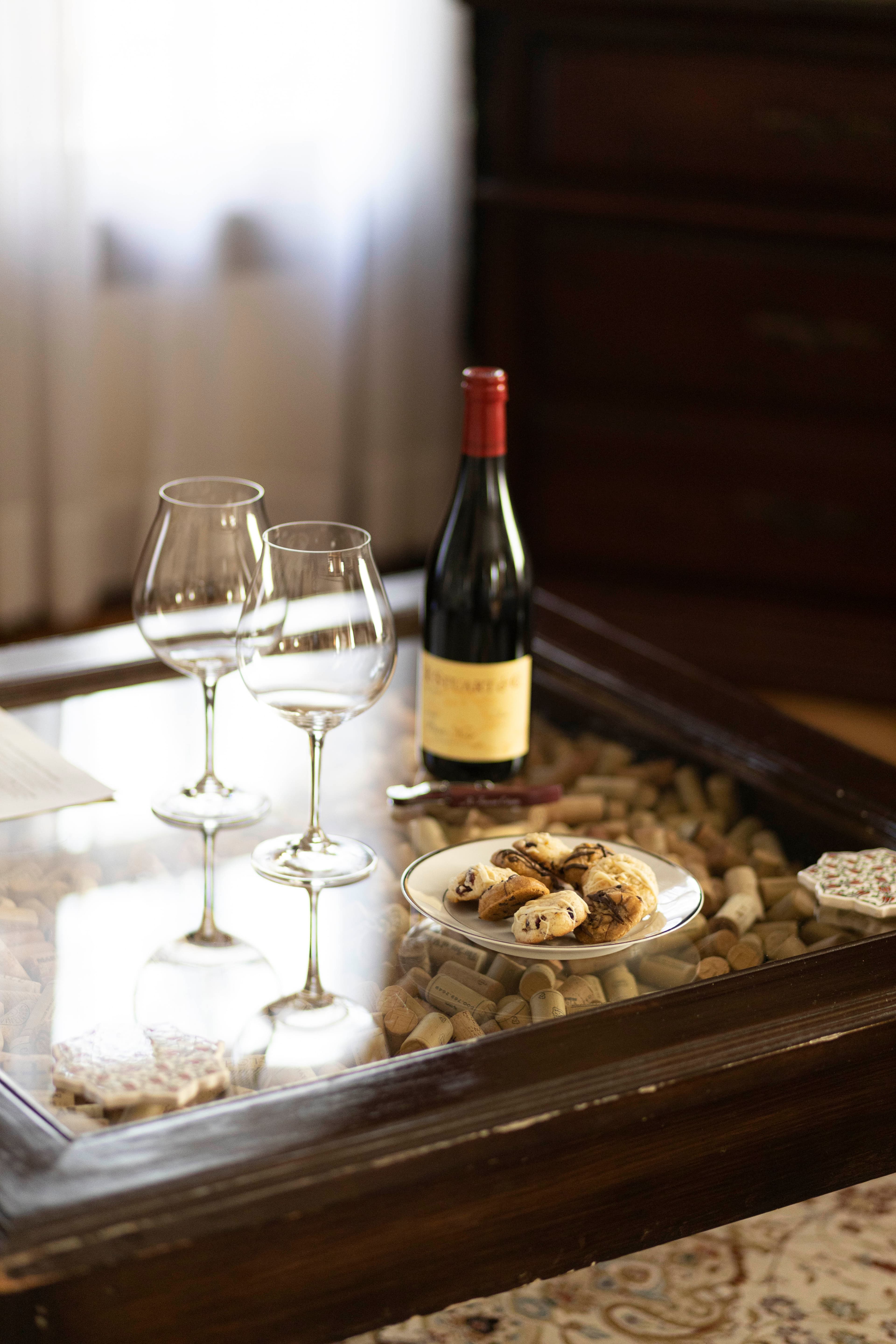 A close-up of a glass-topped table with two wine glasses, a bottle of red wine, and a plate of cookies, set against a blurred background.