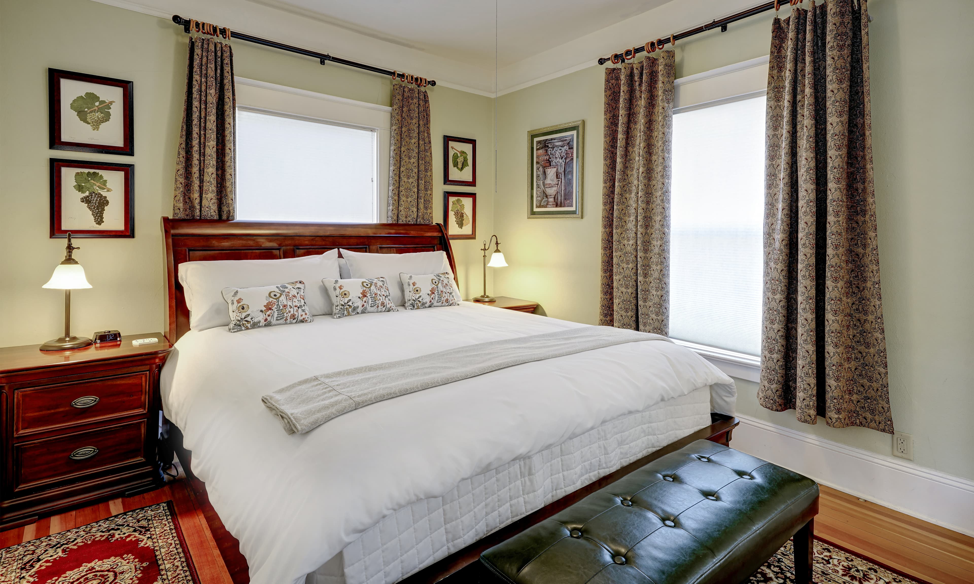 View of California king sleigh bed with two side bureaus and green leather bench as well as two exterior windows.