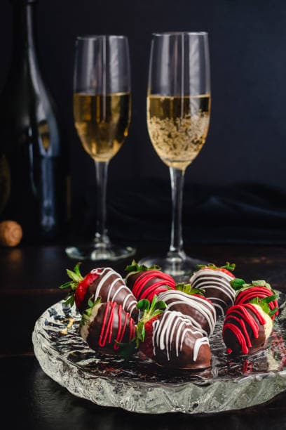 A plate of chocolate-covered strawberries with colorful drizzles, accompanied by two flutes of sparkling wine.