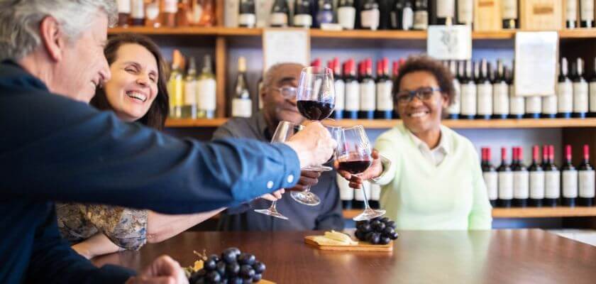 A group of four people toasting with glasses of wine at a table, with shelves of wine bottles in the background.