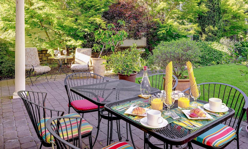 A beautifully arranged outdoor dining space with colorful chairs, a glass table set with breakfast items, and lush greenery in the background.