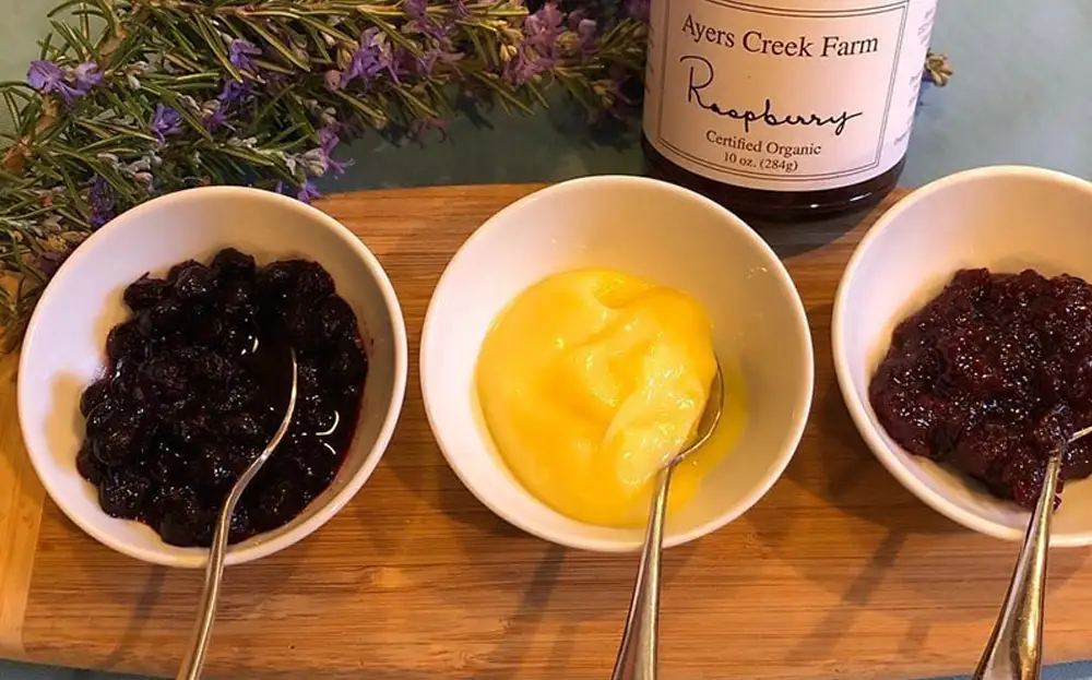 Three bowls containing different flavored preserves-berry, lemon, and raspberry-on a wooden platter, with a jar of raspberry preserve in the background.