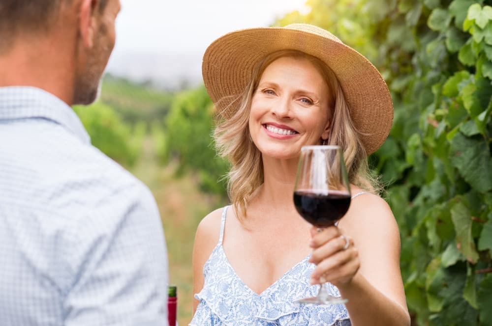 A woman in a straw hat smiles and raises a glass of red wine in a vineyard.