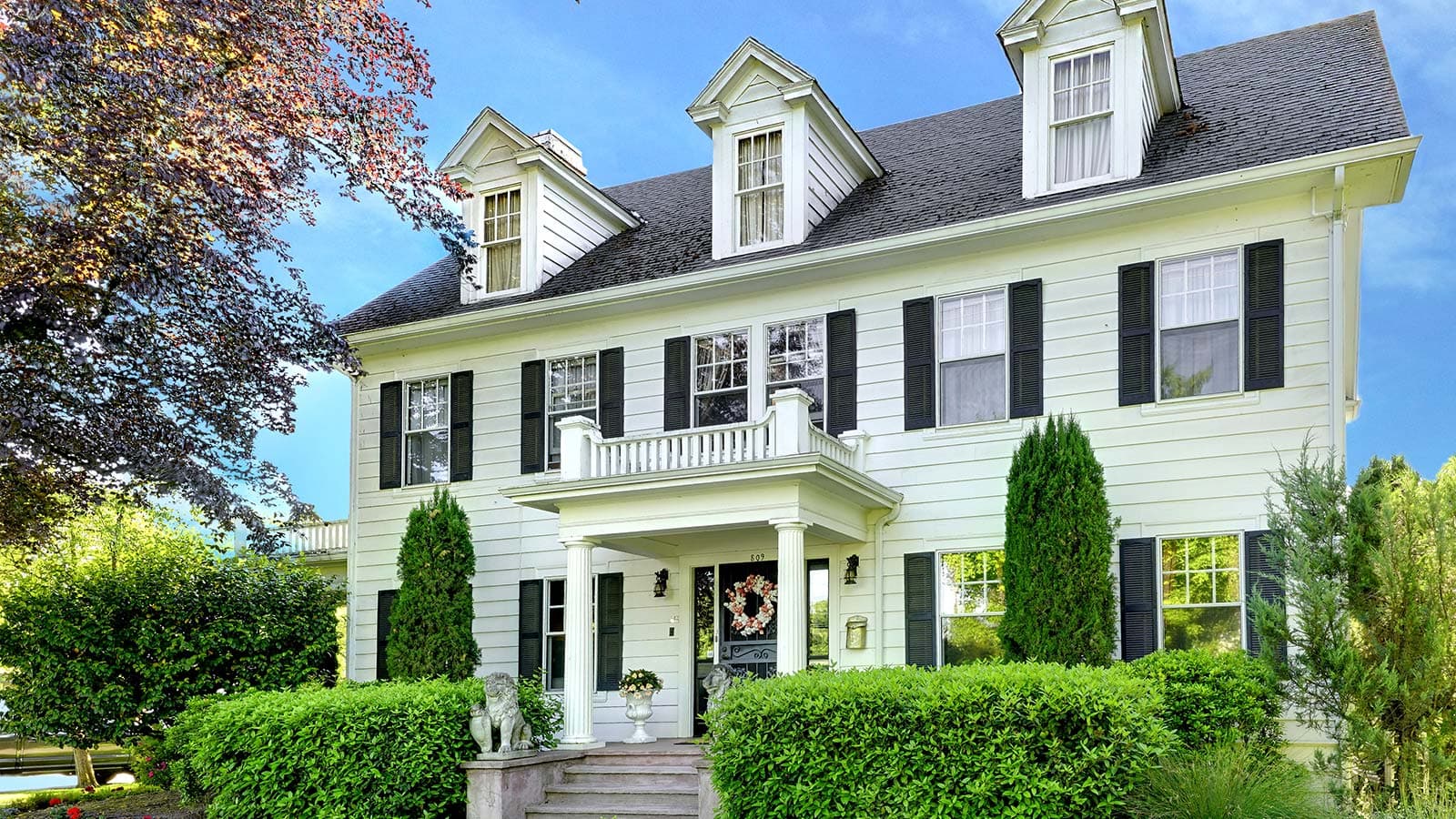 A charming two-story white house with black shutters and a landscaped front garden.