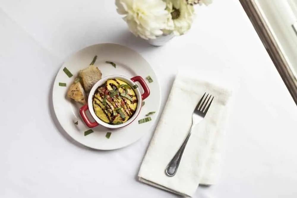 A small red dish filled with a colorful baked dish, accompanied by two pieces of bread, a fork, and a white napkin on a table.
