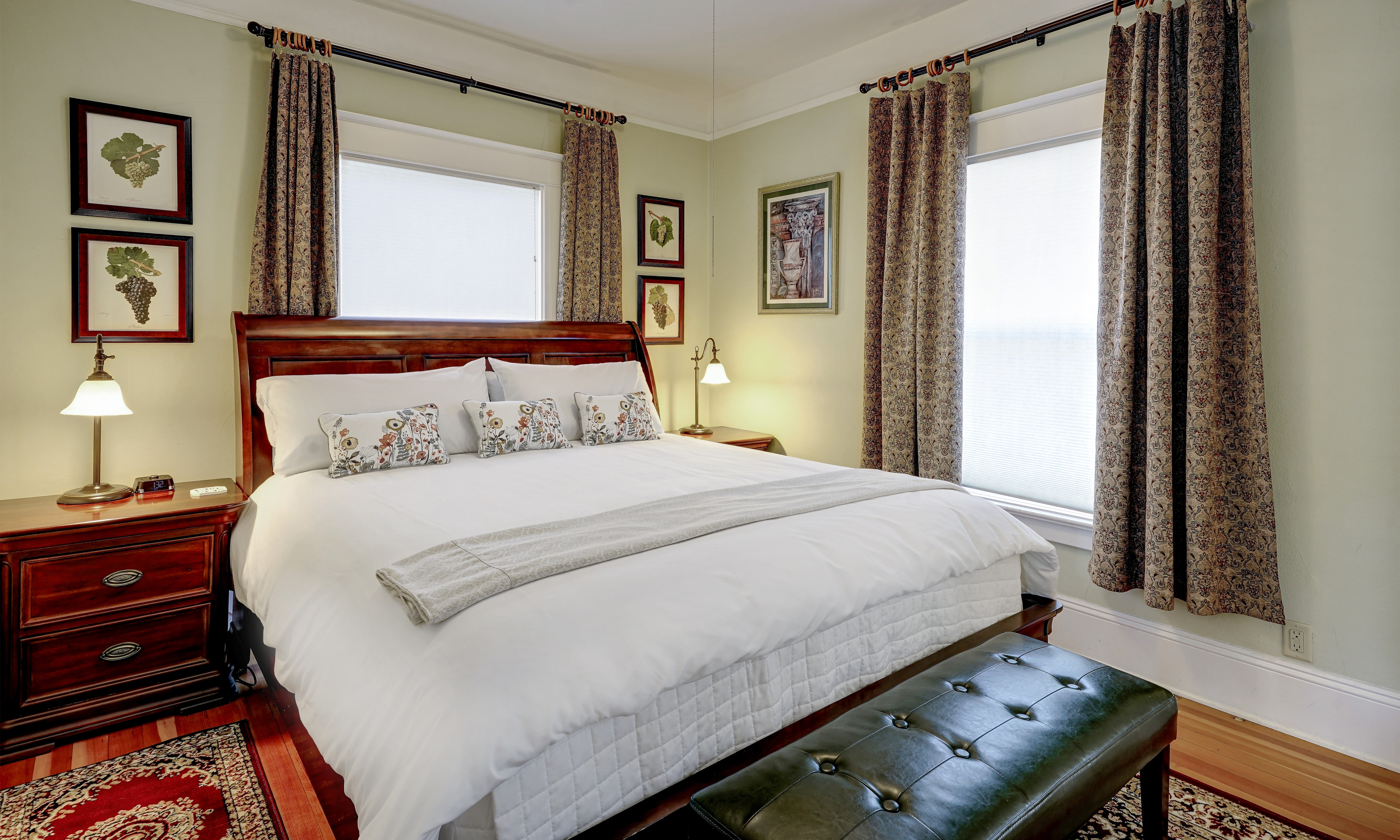 View of California king sleigh bed with two side bureaus and green leather bench as well as two exterior windows.