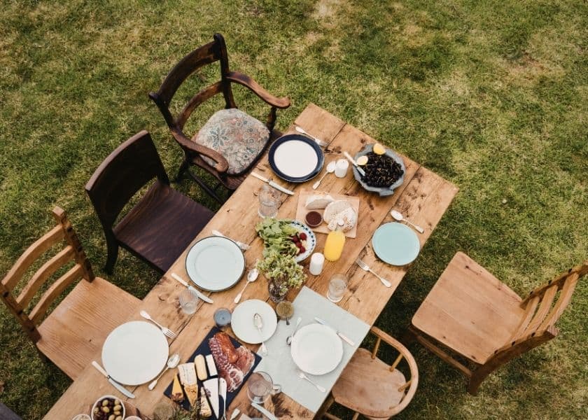 An aerial view of a rustic wooden table set for a meal with empty plates and various food items, surrounded by chairs on green grass. An aerial view of a rustic wooden table set for a meal with empty plates and various food items, surrounded by chairs on green grass.