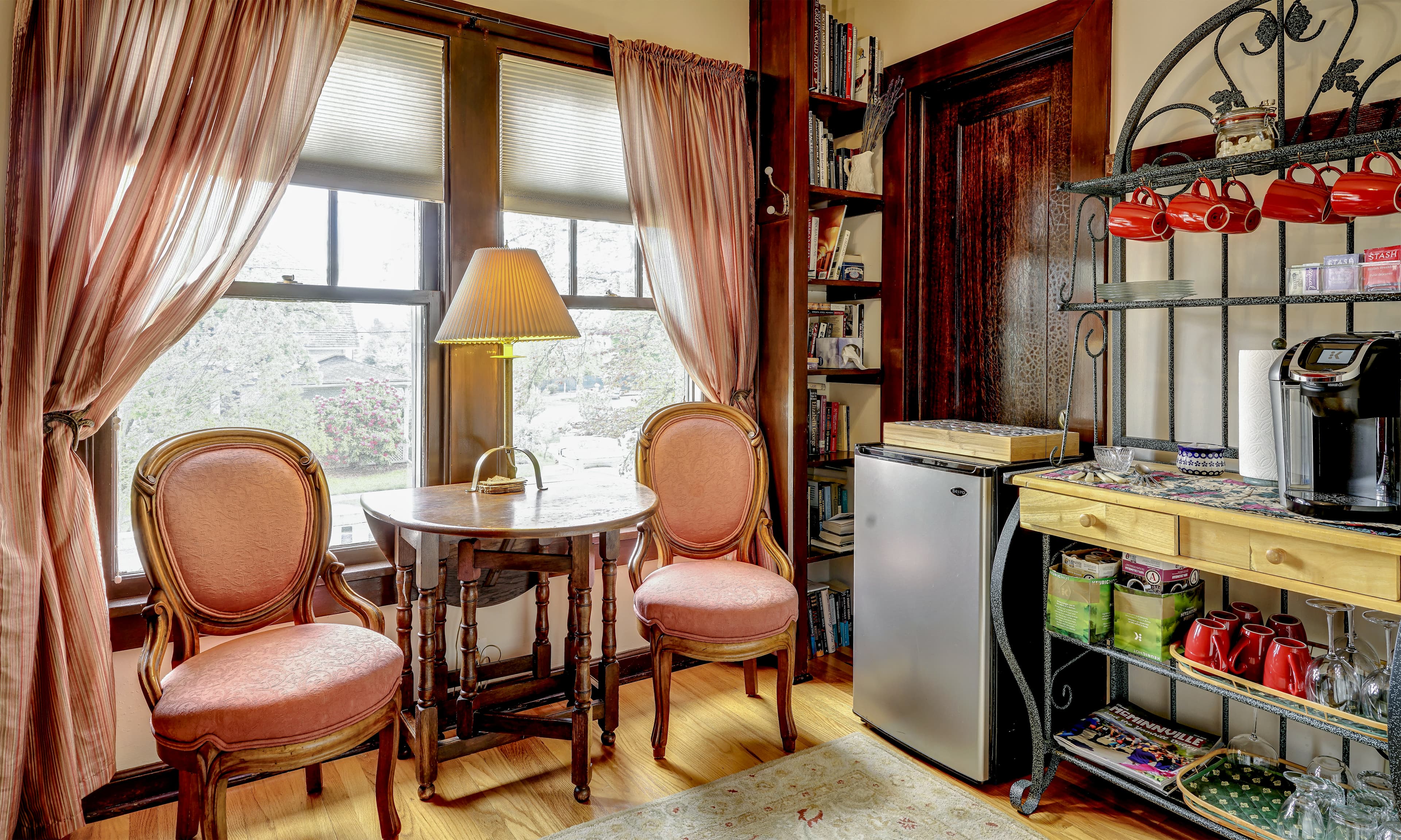 A cozy corner with two pink chairs, a small table, a lamp, and a kitchenette featuring a mini fridge and coffee station.