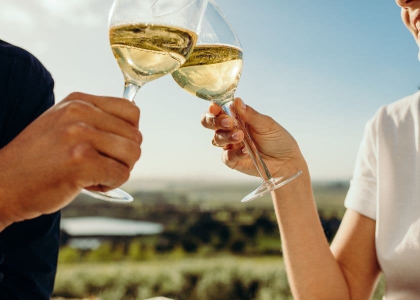 Two hands clink glasses of white wine with a scenic backdrop.