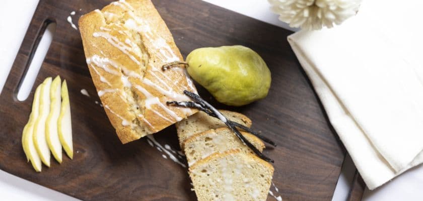 A loaf of vanilla pear cake, sliced and served with a pear and lemon slices on a wooden board.