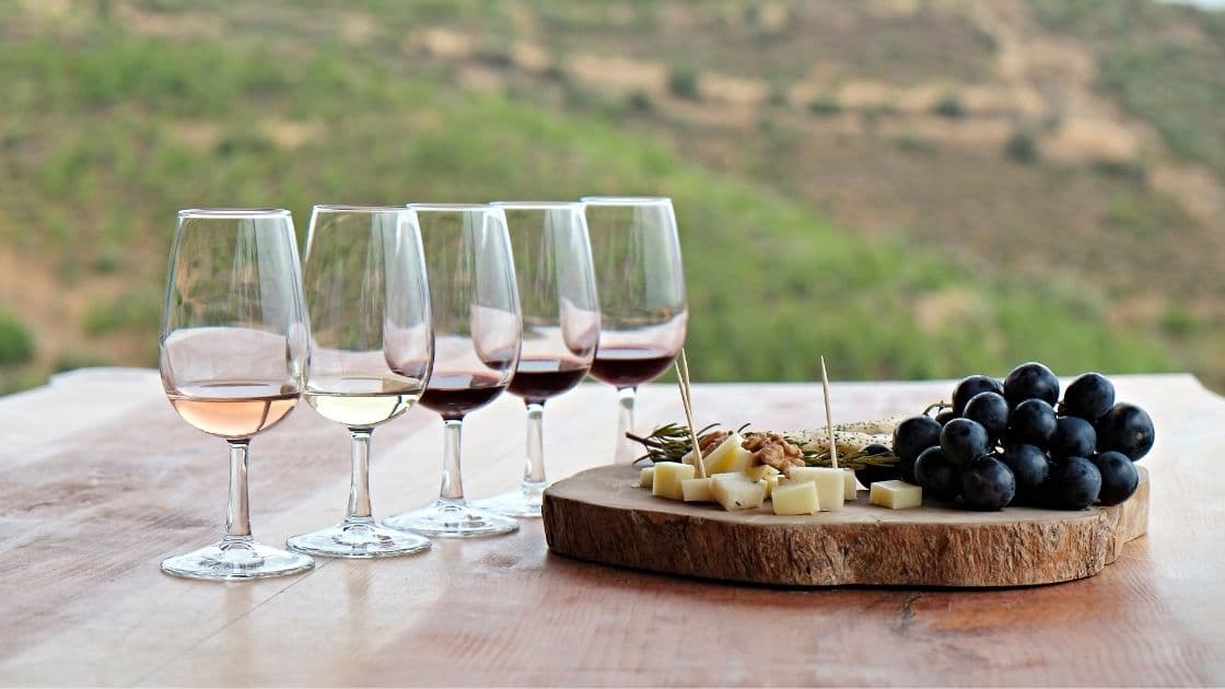 Five wine glasses are lined up beside a wooden platter with cheese, grapes, and snacks, set against a green landscape.