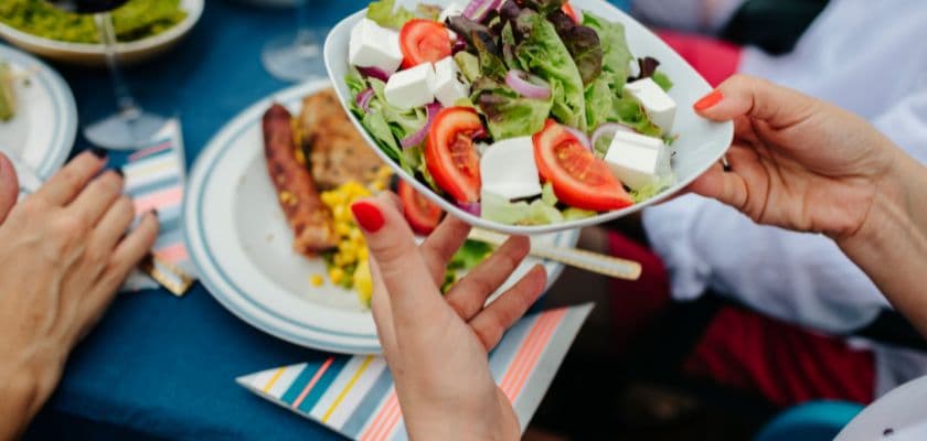 A person hands a fresh salad with tomatoes and feta cheese at a dining table.