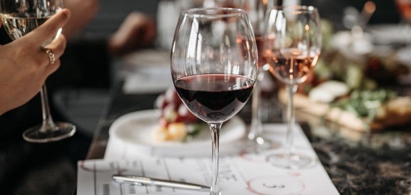 A close-up of a red wine glass at a dining table, surrounded by other glasses and a plate with food.