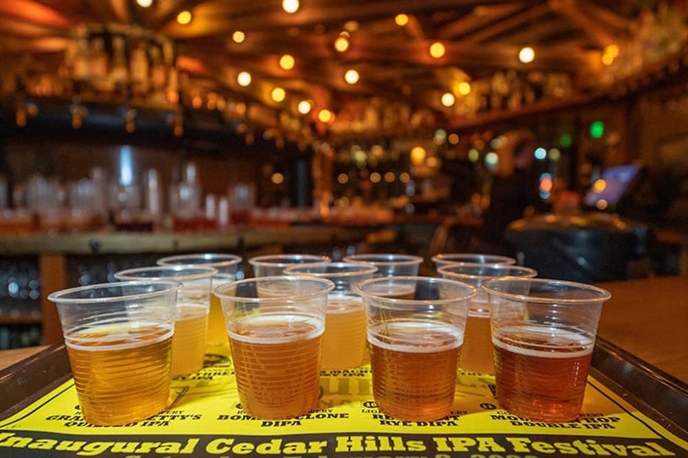 A tray of various craft beers at the Inaugural Cedar Hills IPA Festival, illuminated by warm lights in a bar setting.