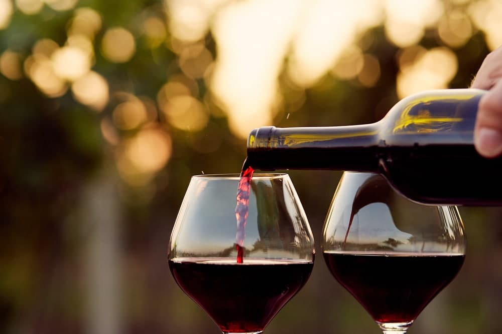 A bottle of red wine is being poured into two glasses against a blurred vineyard background.