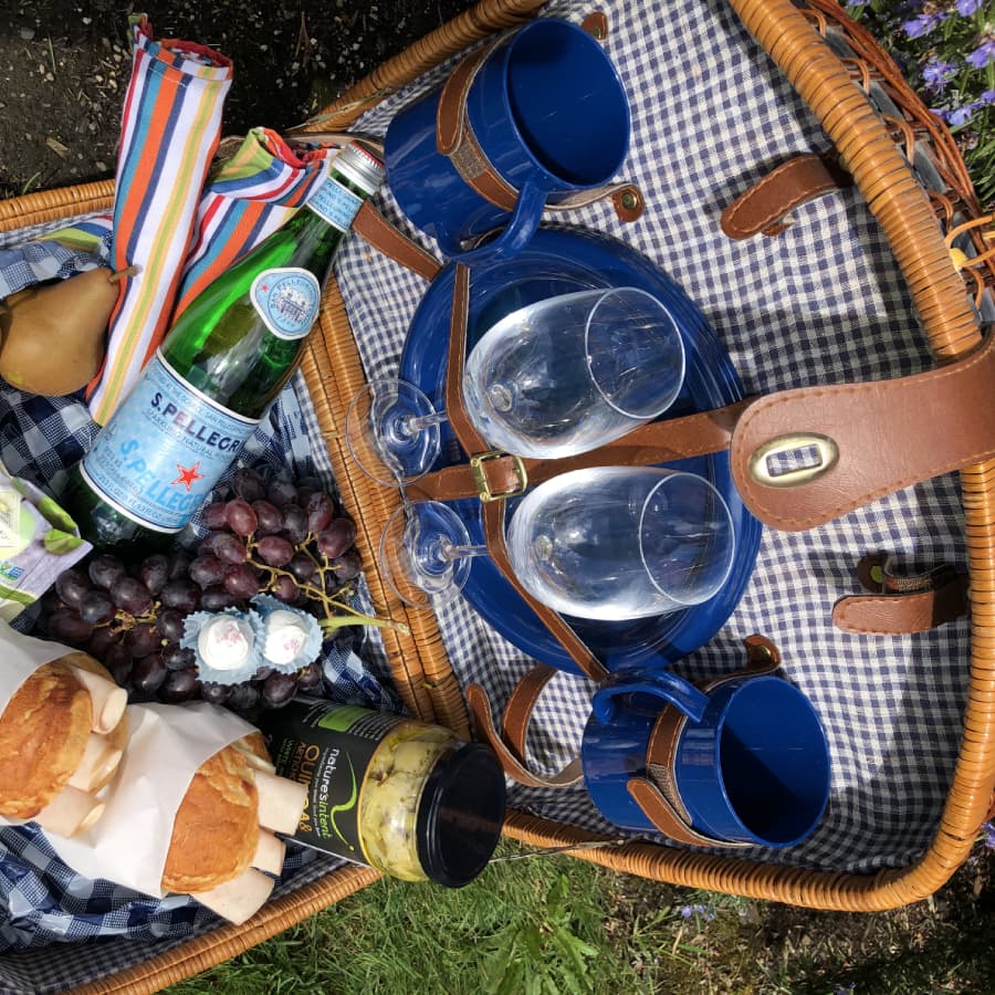 A picnic basket filled with wine glasses, a bottle of sparkling water, grapes, pastries, and utensils on a checkered cloth.
