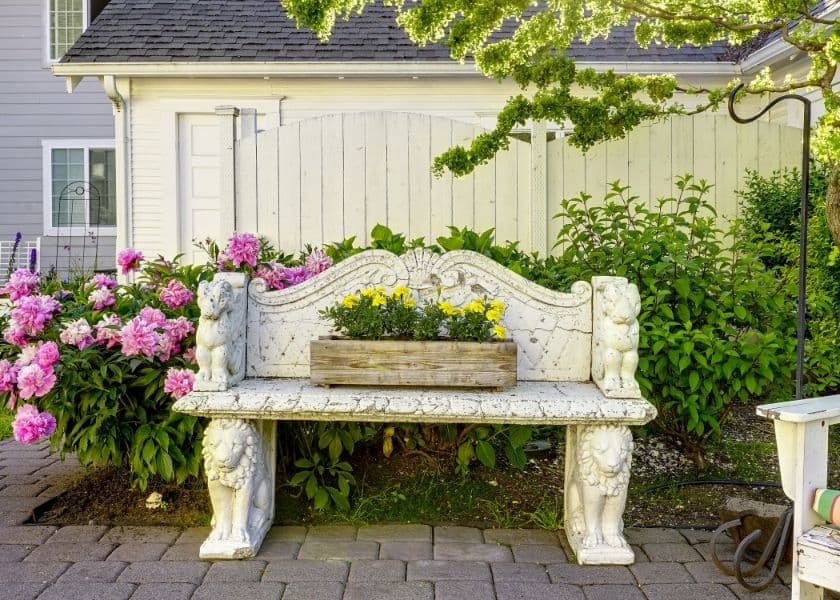 A white, ornate bench adorned with lion-shaped ends sits amid vibrant pink flowers and greenery. A white, ornate bench adorned with lion-shaped ends sits amid vibrant pink flowers and greenery.